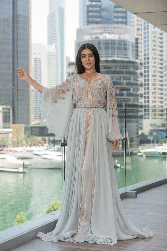 Femme portant un caftan marocain élégant en couleur douce, avec des broderies intricées, debout sur un balcon surplombant un port moderne et un paysage urbain.